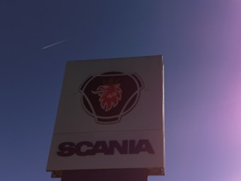 A tall sign displays a logo featuring a red griffin's head within a shield-like emblem, above the prominent text 'SCANIA'. The background is a clear blue sky with a faint jet contrail visible.
