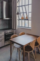 A modern kitchen setting features a wooden dining table with four matching chairs, adjacent to a large window with black frames. The ceiling has pendant lights hanging above the table. The kitchen cabinetry is sleek, with a black cooktop and stainless steel vent hood. Neutral tones and natural light create a cozy and inviting atmosphere.