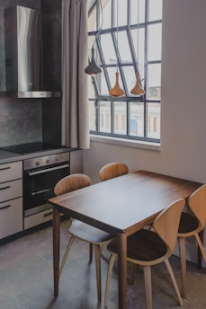 A modern kitchen setting features a wooden dining table with four matching chairs, adjacent to a large window with black frames. The ceiling has pendant lights hanging above the table. The kitchen cabinetry is sleek, with a black cooktop and stainless steel vent hood. Neutral tones and natural light create a cozy and inviting atmosphere.