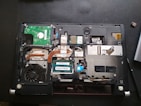A laptop with its back cover removed, revealing various internal components including a cooling fan, circuit boards, copper heat pipes, and RAM modules. The layout shows intricate electronic parts and connectors.