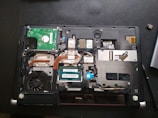 A laptop with its back cover removed, revealing various internal components including a cooling fan, circuit boards, copper heat pipes, and RAM modules. The layout shows intricate electronic parts and connectors.