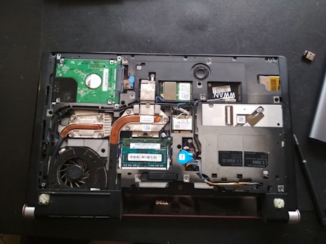 A laptop with its back cover removed, revealing various internal components including a cooling fan, circuit boards, copper heat pipes, and RAM modules. The layout shows intricate electronic parts and connectors.