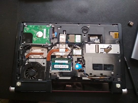 A laptop with its back cover removed, revealing various internal components including a cooling fan, circuit boards, copper heat pipes, and RAM modules. The layout shows intricate electronic parts and connectors.
