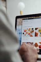 A person is using a laptop, navigating through the Uber Eats food delivery website. The screen displays various food items available for delivery in Montreal, QC, including options like McDonald's and Thai Express. The person appears to be browsing with their hand near the trackpad.