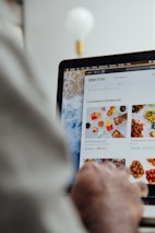 A person is using a laptop, navigating through the Uber Eats food delivery website. The screen displays various food items available for delivery in Montreal, QC, including options like McDonald's and Thai Express. The person appears to be browsing with their hand near the trackpad.