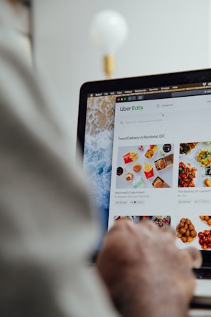 A person is using a laptop, navigating through the Uber Eats food delivery website. The screen displays various food items available for delivery in Montreal, QC, including options like McDonald's and Thai Express. The person appears to be browsing with their hand near the trackpad.