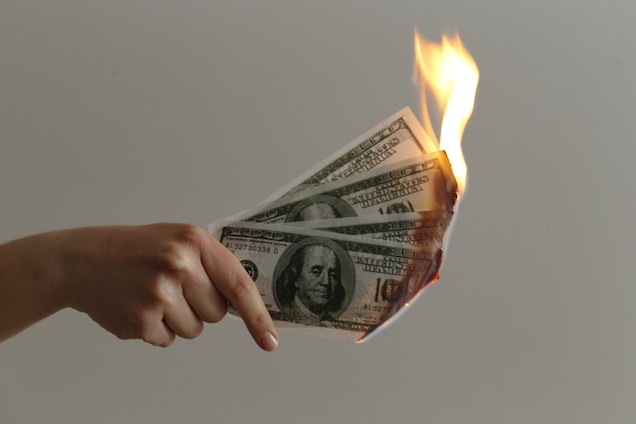 A hand holding several burning US dollar bills with flames visible at one end. The background is a plain, neutral color.