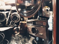 A vintage camera collection featuring an Instamatic camera and various other antique cameras and accessories, placed on a reflective surface. The image has a warm, nostalgic feel with soft lighting highlighting the metal and glass components.