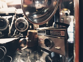 A vintage camera collection featuring an Instamatic camera and various other antique cameras and accessories, placed on a reflective surface. The image has a warm, nostalgic feel with soft lighting highlighting the metal and glass components.