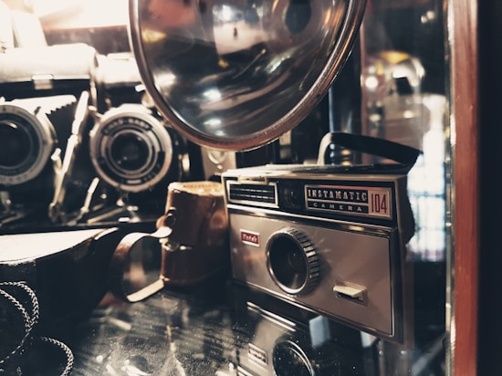 A vintage camera collection featuring an Instamatic camera and various other antique cameras and accessories, placed on a reflective surface. The image has a warm, nostalgic feel with soft lighting highlighting the metal and glass components.