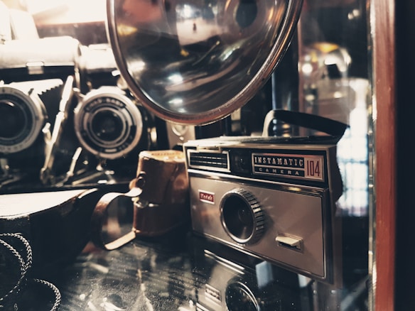 A vintage camera collection featuring an Instamatic camera and various other antique cameras and accessories, placed on a reflective surface. The image has a warm, nostalgic feel with soft lighting highlighting the metal and glass components.