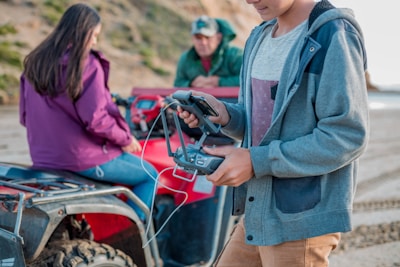 A person in a gray hoodie operates a remote control for a drone while standing next to an ATV. Another individual sits on the ATV, facing away, wearing a purple jacket. A third person in the background, wearing a dark green jacket and cap, leans on the vehicle looking down. The setting appears to be outdoors, possibly on a beach or near a cliffside.