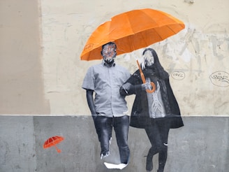 A piece of street art on a wall depicts two figures standing together under an orange umbrella. The faces of the figures are obscured or non-detailed, adding an abstract element to the image. The wall behind them is lightly textured and features various graffiti markings. A smaller, painted orange umbrella appears on the wall beside them.