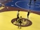 Two wrestlers and a referee are on a yellow wrestling mat with a maroon and gold logo in the center. One wrestler is wearing a black singlet with a gear headguard, and the other wrestler is also in a black singlet appearing ready to start the match. The referee stands between them, observing the scene. An adjacent wrestling mat is visible in the background, colored in blue, where another pair of wrestlers seem to be competing.