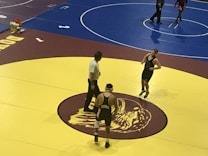 Two wrestlers and a referee are on a yellow wrestling mat with a maroon and gold logo in the center. One wrestler is wearing a black singlet with a gear headguard, and the other wrestler is also in a black singlet appearing ready to start the match. The referee stands between them, observing the scene. An adjacent wrestling mat is visible in the background, colored in blue, where another pair of wrestlers seem to be competing.