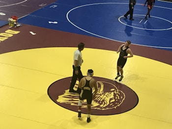 Two wrestlers and a referee are on a yellow wrestling mat with a maroon and gold logo in the center. One wrestler is wearing a black singlet with a gear headguard, and the other wrestler is also in a black singlet appearing ready to start the match. The referee stands between them, observing the scene. An adjacent wrestling mat is visible in the background, colored in blue, where another pair of wrestlers seem to be competing.
