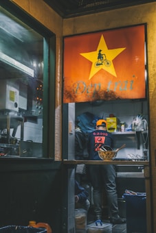 A person wearing an orange cap is working in a small, dimly lit kitchen. The interior has various kitchen items and ingredients on shelves, with a bowl and utensils on the counter. The entrance features a large red sign with a yellow star and the text 'Đẹp trai'.