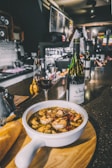 A bowl of seafood soup with shrimp and other ingredients is placed on a wooden board, accompanied by a piece of bread. A glass of red wine and a bottle are set next to the soup on a dark, shiny countertop with a bar in the background, creating an inviting atmosphere.