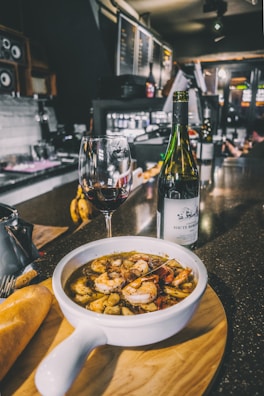 A bowl of seafood soup with shrimp and other ingredients is placed on a wooden board, accompanied by a piece of bread. A glass of red wine and a bottle are set next to the soup on a dark, shiny countertop with a bar in the background, creating an inviting atmosphere.