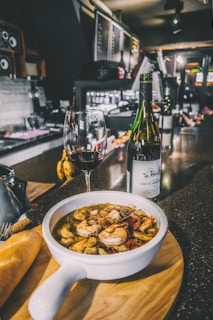 A bowl of seafood soup with shrimp and other ingredients is placed on a wooden board, accompanied by a piece of bread. A glass of red wine and a bottle are set next to the soup on a dark, shiny countertop with a bar in the background, creating an inviting atmosphere.