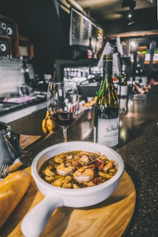 A bowl of seafood soup with shrimp and other ingredients is placed on a wooden board, accompanied by a piece of bread. A glass of red wine and a bottle are set next to the soup on a dark, shiny countertop with a bar in the background, creating an inviting atmosphere.