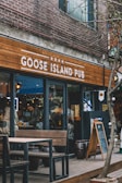 A pub with a rustic wooden sign reading 'Goose Island Pub'. The exterior features brick walls and glass doors. There are outdoor wooden tables and benches. Inside, people are interacting, adding a lively atmosphere. The space is decorated with lights and various wooden elements.