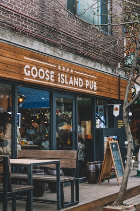 A pub with a rustic wooden sign reading 'Goose Island Pub'. The exterior features brick walls and glass doors. There are outdoor wooden tables and benches. Inside, people are interacting, adding a lively atmosphere. The space is decorated with lights and various wooden elements.