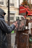 An artist wearing a hooded jacket is painting on a canvas set up on an easel in an outdoor setting. The scene depicts a street or market area, with a red awning and a Christmas tree with lights in the background, contributing to a festive atmosphere.