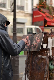 An artist wearing a hooded jacket is painting on a canvas set up on an easel in an outdoor setting. The scene depicts a street or market area, with a red awning and a Christmas tree with lights in the background, contributing to a festive atmosphere.