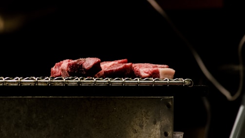 Slices of raw meat rest on a metallic grill with a black background, highlighting the texture and marbling of the meat.