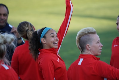 Several individuals wearing red athletic tracksuits are gathered outdoors. One person with a ponytail and headband is joyfully raising an arm. Another person with a shaved side hairstyle is also visible, engaging in the group interaction.