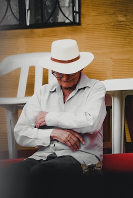 An elderly person wearing a white hat and dress shirt is seated with arms crossed. The setting includes white plastic chairs and a yellowish textured wall, with a small cat resting at the person's feet.