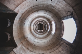 A view looking upward through the interior of a large, cylindrical concrete structure with a series of concentric rings. The structure appears weathered, with light coming from gaps and openings above.