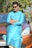 A man wearing sunglasses and a blue traditional kurta is leaning against the front of an orange car. The scene is outdoors with trees in the background and a road nearby.