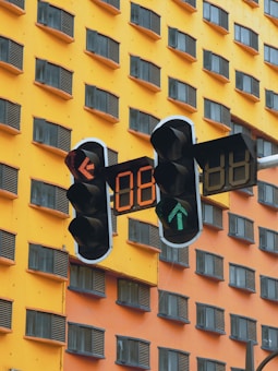 A traffic light with digital countdown timers is positioned in front of a tall building. The building has a vibrant yellow and orange facade with numerous windows featuring dark shutters. The traffic light displays a green arrow and red numbers changing.