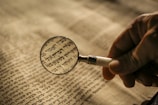 A hand holding a magnifying glass over a text written in Hebrew. The focus is on a specific section, enlarging the words under the convex lens. The paper appears aged, with a warm tone, suggesting an ancient or religious manuscript.
