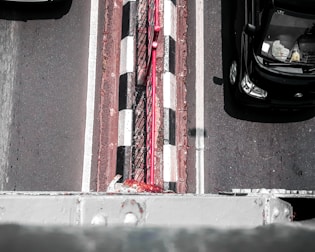 An aerial view of a road with two visible lanes and a narrow pedestrian strip in the center. The road has black and white checkered markings and a red railing running alongside the pedestrian strip. A black car is traveling in one lane, partially visible with objects seen inside through the windshield.