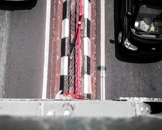 An aerial view of a road with two visible lanes and a narrow pedestrian strip in the center. The road has black and white checkered markings and a red railing running alongside the pedestrian strip. A black car is traveling in one lane, partially visible with objects seen inside through the windshield.
