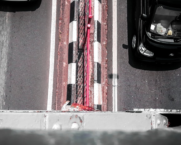 An aerial view of a road with two visible lanes and a narrow pedestrian strip in the center. The road has black and white checkered markings and a red railing running alongside the pedestrian strip. A black car is traveling in one lane, partially visible with objects seen inside through the windshield.