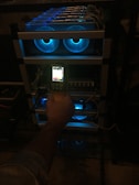 A hand holding a thermal scanner is pointed towards a rack of computer servers. The servers are illuminated with blue LED lights, giving a technological and futuristic feel.