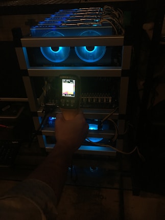 A hand holding a thermal scanner is pointed towards a rack of computer servers. The servers are illuminated with blue LED lights, giving a technological and futuristic feel.