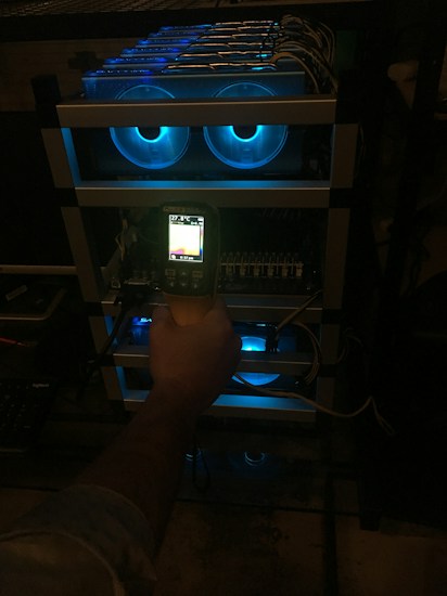 A hand holding a thermal scanner is pointed towards a rack of computer servers. The servers are illuminated with blue LED lights, giving a technological and futuristic feel.