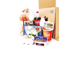 A collection of various grocery items displayed together. There is a loaf of bread in a bag, a plastic bottle of Coca-Cola, a carton of eggs, milk, yogurt containers, a packet of butter, and a plastic tray of vegetables. Behind these items, there is a cardboard box. The products feature labels in a non-English script.
