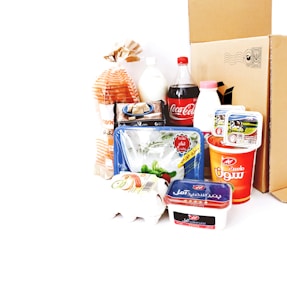 A collection of various grocery items displayed together. There is a loaf of bread in a bag, a plastic bottle of Coca-Cola, a carton of eggs, milk, yogurt containers, a packet of butter, and a plastic tray of vegetables. Behind these items, there is a cardboard box. The products feature labels in a non-English script.