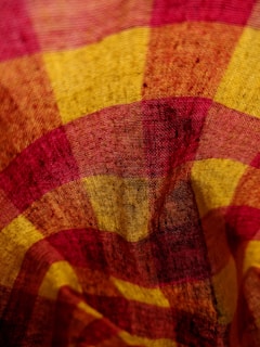 A close-up view of a textured fabric with a pattern of intersecting lines, primarily in shades of red and yellow, creating a plaid-like appearance. The material has a slightly wrinkled texture, emphasizing its weave.
