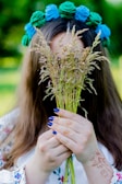 A person holds a bouquet of wild grasses, partially obscuring their face. They have long hair and wear a floral headdress with blue and green flowers. Their hands display intricate henna designs and they have blue painted nails. The setting appears to be outdoors with blurred greenery in the background.