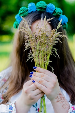 A person holds a bouquet of wild grasses, partially obscuring their face. They have long hair and wear a floral headdress with blue and green flowers. Their hands display intricate henna designs and they have blue painted nails. The setting appears to be outdoors with blurred greenery in the background.
