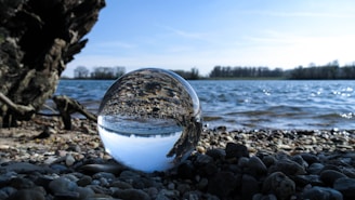 A crystal ball sits on a pebble-covered shore, reflecting an inverted view of a calm lake and the sky. The background features a clear blue sky, distant trees lining the horizon, and gentle waves rippling across the water surface.