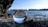 A crystal ball sits on a pebble-covered shore, reflecting an inverted view of a calm lake and the sky. The background features a clear blue sky, distant trees lining the horizon, and gentle waves rippling across the water surface.