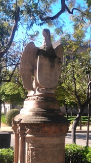 A graceful outdoor statue of a bird perched on a rustic pedestal in a sunlit garden.